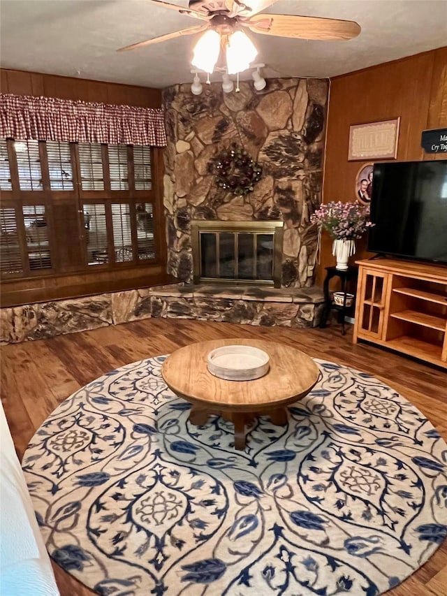 living area with wood-type flooring, wooden walls, ceiling fan, and a stone fireplace