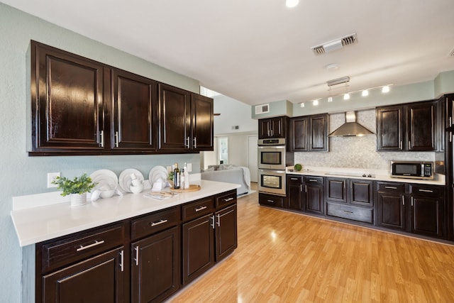 kitchen featuring light hardwood / wood-style floors, stainless steel appliances, dark brown cabinetry, wall chimney range hood, and tasteful backsplash