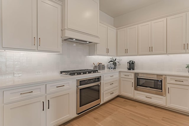 kitchen featuring stainless steel appliances, white cabinetry, custom range hood, light hardwood / wood-style floors, and backsplash