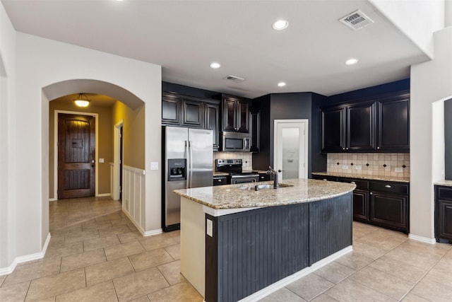 kitchen with light stone countertops, stainless steel appliances, an island with sink, decorative backsplash, and sink