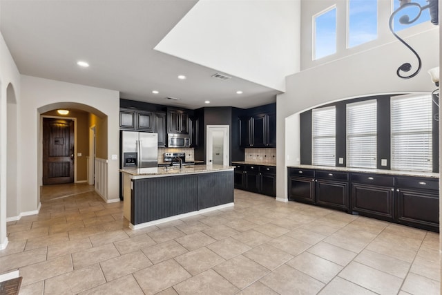 kitchen featuring a kitchen island with sink, stainless steel appliances, light stone countertops, decorative backsplash, and light tile patterned flooring