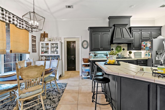 kitchen with tile counters, light tile patterned floors, a notable chandelier, stainless steel fridge, and ornamental molding