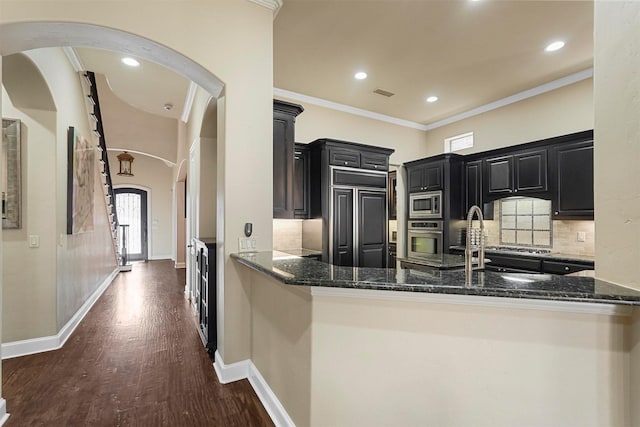 kitchen featuring kitchen peninsula, dark stone countertops, built in appliances, and ornamental molding