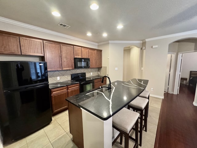 kitchen with sink, light tile patterned floors, decorative backsplash, a center island with sink, and black appliances