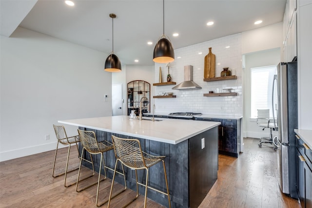 kitchen featuring stainless steel fridge, hanging light fixtures, wall chimney range hood, and a center island with sink