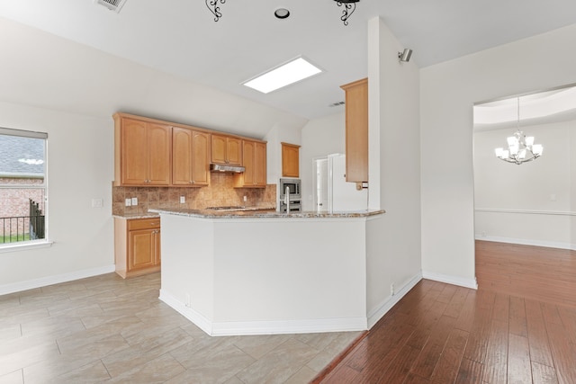 kitchen with light stone countertops, kitchen peninsula, a notable chandelier, decorative backsplash, and stainless steel microwave