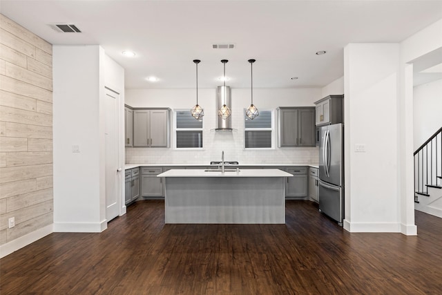 kitchen featuring wood walls, stainless steel refrigerator, hanging light fixtures, an island with sink, and wall chimney exhaust hood