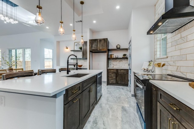 kitchen with an island with sink, wall chimney range hood, backsplash, electric range oven, and sink