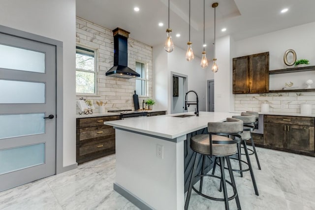 kitchen featuring pendant lighting, wall chimney range hood, a kitchen island with sink, dark brown cabinets, and sink