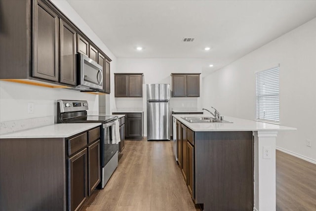 kitchen with sink, light hardwood / wood-style floors, a kitchen island with sink, appliances with stainless steel finishes, and dark brown cabinetry