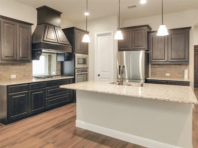 kitchen with stainless steel appliances, premium range hood, a kitchen island with sink, and decorative backsplash