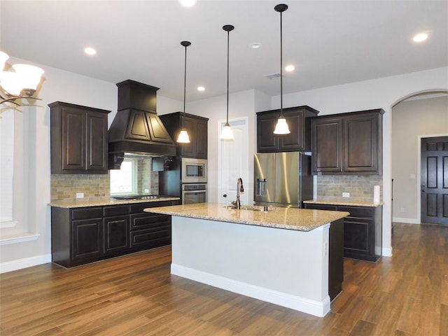 kitchen with stainless steel appliances, decorative light fixtures, tasteful backsplash, custom exhaust hood, and a center island with sink