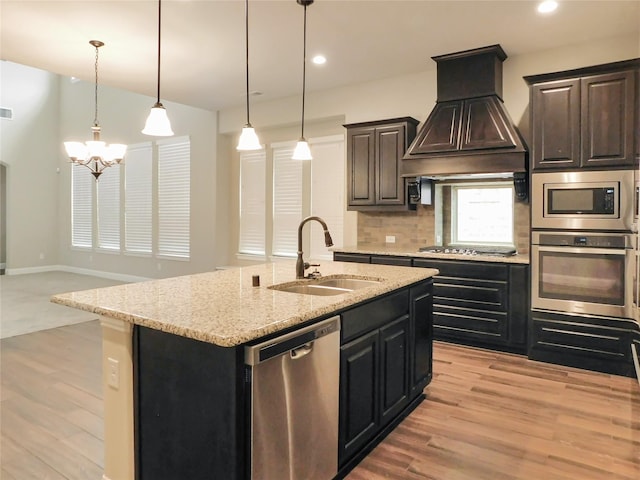 kitchen featuring stainless steel appliances, sink, ventilation hood, a center island with sink, and pendant lighting