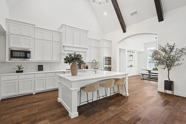 kitchen featuring built in microwave, a center island with sink, white cabinets, and beamed ceiling