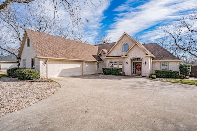 french country home with a garage, concrete driveway, brick siding, and roof with shingles