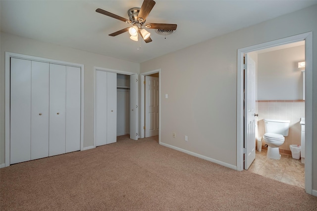 unfurnished bedroom featuring two closets, connected bathroom, tile walls, ceiling fan, and light colored carpet