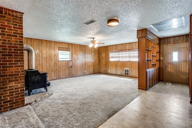 unfurnished living room featuring wooden walls, ceiling fan, a wood stove, and light colored carpet