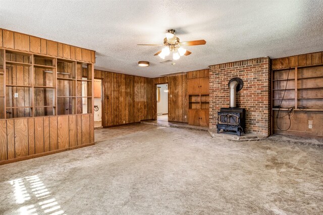 unfurnished living room with wooden walls, a textured ceiling, a wood stove, light carpet, and ceiling fan