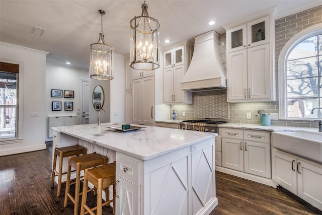 kitchen with light stone countertops, premium range hood, decorative light fixtures, an island with sink, and sink