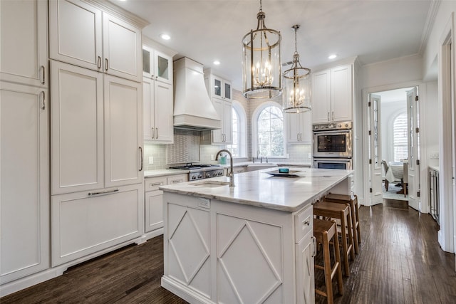 kitchen with custom range hood, double oven, white cabinets, and an island with sink