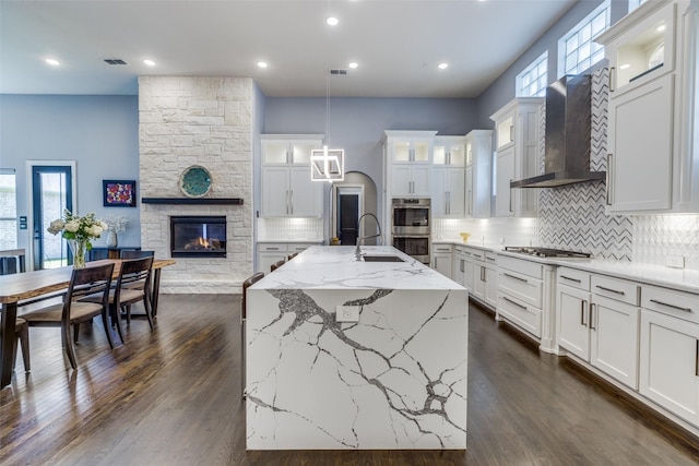 kitchen with a stone fireplace, a kitchen island with sink, sink, white cabinetry, and wall chimney range hood