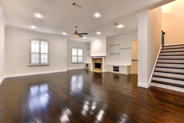 unfurnished living room with a tiled fireplace, crown molding, ceiling fan, dark wood-type flooring, and built in shelves