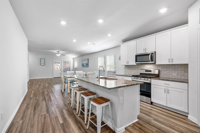 kitchen with stainless steel appliances, a center island with sink, dark stone countertops, white cabinets, and ceiling fan