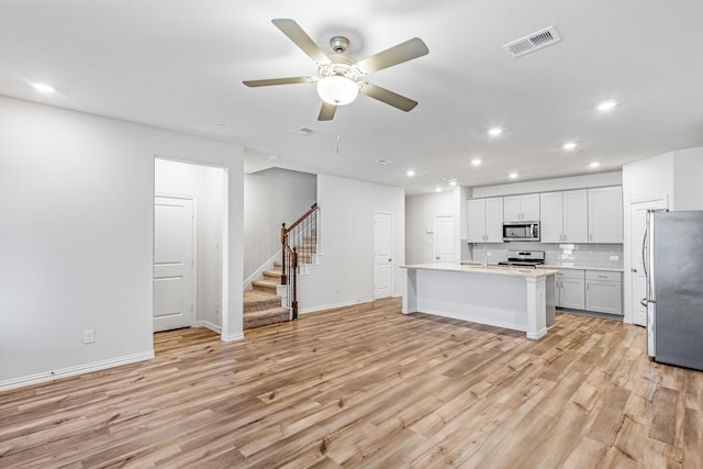 kitchen with a center island with sink, decorative backsplash, light wood-type flooring, appliances with stainless steel finishes, and ceiling fan