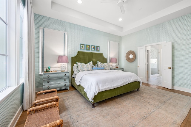 bedroom featuring ceiling fan, ensuite bath, hardwood / wood-style flooring, and a raised ceiling