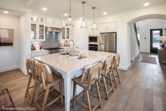 kitchen with a kitchen island with sink, appliances with stainless steel finishes, hanging light fixtures, white cabinetry, and exhaust hood