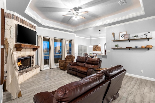 living room with crown molding, a large fireplace, a raised ceiling, ceiling fan, and light hardwood / wood-style floors