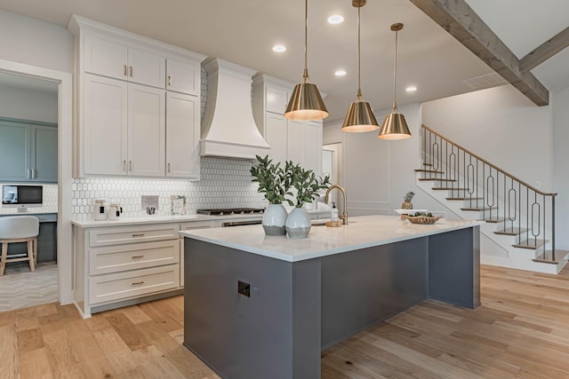 kitchen featuring white cabinetry, beamed ceiling, hanging light fixtures, premium range hood, and a center island with sink