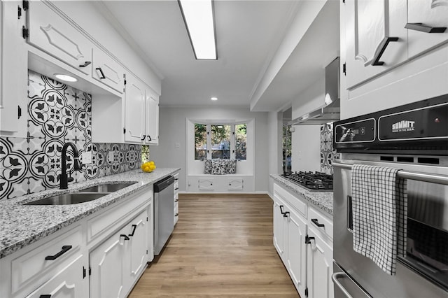 kitchen with light stone counters, stainless steel appliances, wall chimney range hood, white cabinets, and sink