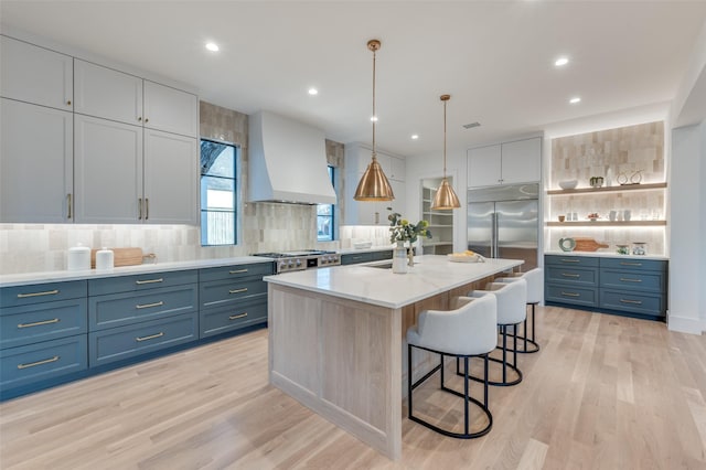 kitchen featuring a kitchen island with sink, hanging light fixtures, built in refrigerator, custom range hood, and light hardwood / wood-style flooring