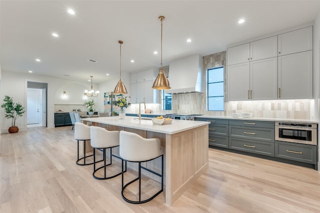 kitchen featuring decorative light fixtures, a center island with sink, gray cabinetry, wall chimney range hood, and backsplash