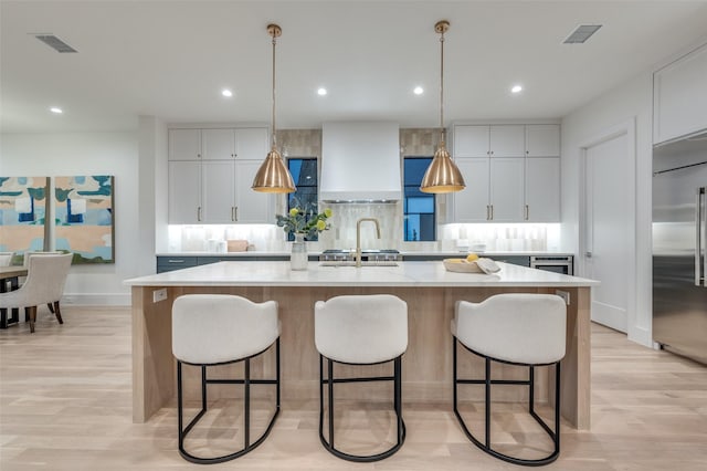 kitchen with stainless steel built in fridge, wall chimney range hood, white cabinetry, and an island with sink