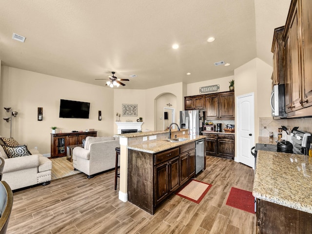 kitchen featuring stainless steel appliances, sink, light stone counters, ceiling fan, and a kitchen island with sink