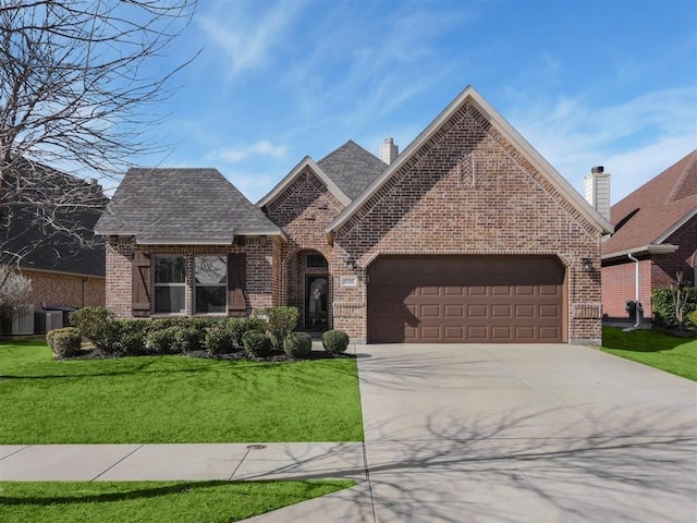 french country inspired facade with a garage, a shingled roof, concrete driveway, a front lawn, and brick siding