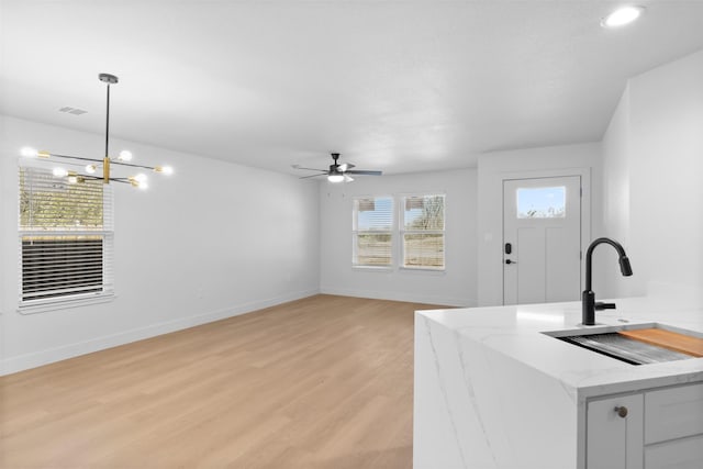 kitchen with sink, light wood-type flooring, ceiling fan with notable chandelier, hanging light fixtures, and light stone countertops