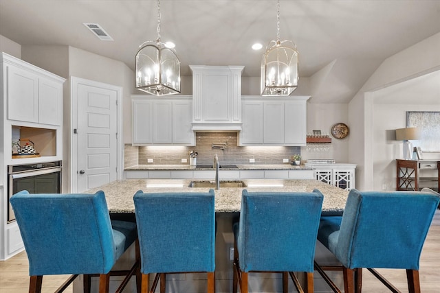 kitchen featuring an island with sink, a notable chandelier, oven, white cabinetry, and decorative light fixtures