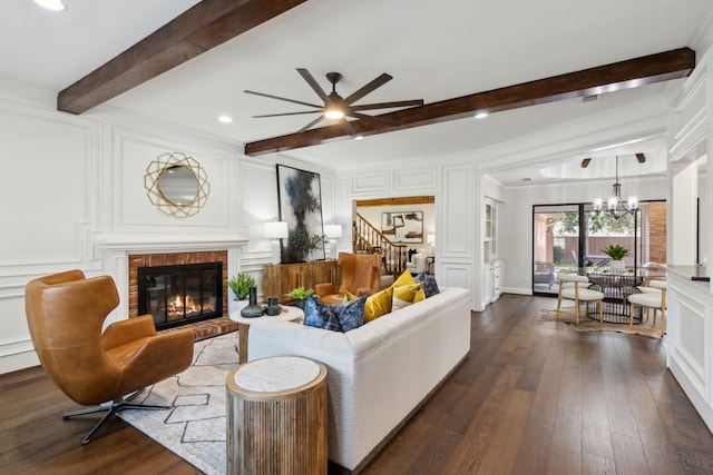 living room featuring dark wood-type flooring, a brick fireplace, ceiling fan with notable chandelier, and beamed ceiling