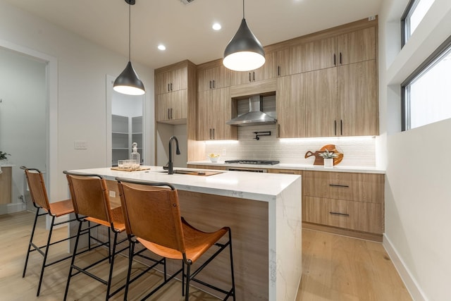 kitchen with a center island with sink, wall chimney exhaust hood, light brown cabinets, and hanging light fixtures