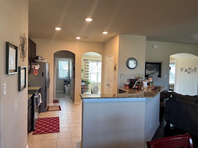 kitchen with kitchen peninsula, dark brown cabinets, stainless steel appliances, light tile patterned floors, and dark stone counters