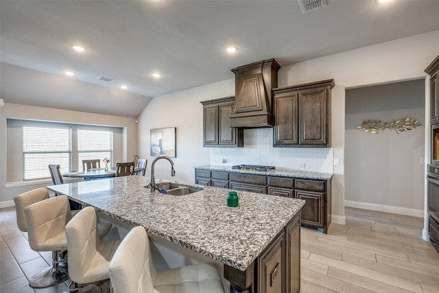 kitchen featuring lofted ceiling, sink, dark brown cabinetry, light stone countertops, and an island with sink