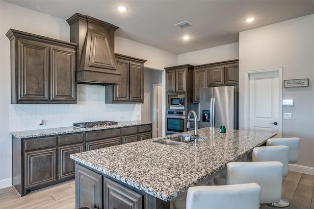 kitchen with stainless steel appliances, premium range hood, sink, a kitchen island with sink, and a breakfast bar