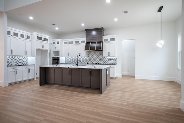 kitchen featuring an island with sink, white cabinets, decorative light fixtures, stainless steel oven, and light wood-type flooring