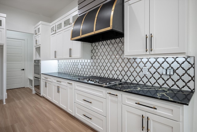 kitchen with exhaust hood, stainless steel gas cooktop, white cabinetry, glass insert cabinets, and light wood-style floors