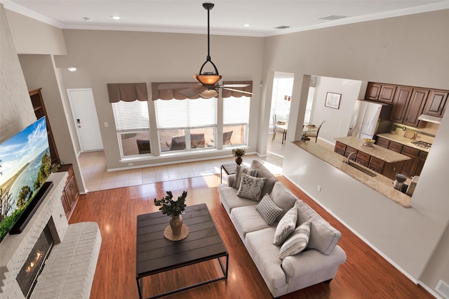 living area with crown molding, light wood finished floors, visible vents, a high ceiling, and a brick fireplace