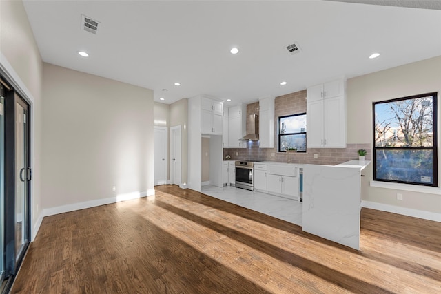 kitchen with electric range, light stone countertops, white cabinetry, wall chimney range hood, and backsplash