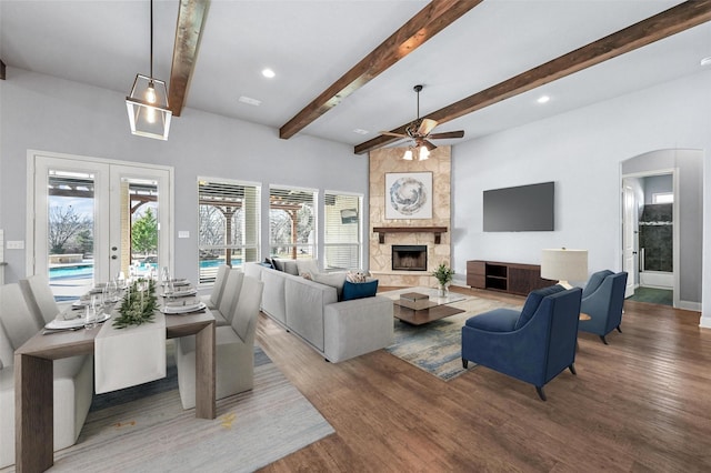 living room featuring ceiling fan, beam ceiling, dark wood-type flooring, a stone fireplace, and french doors